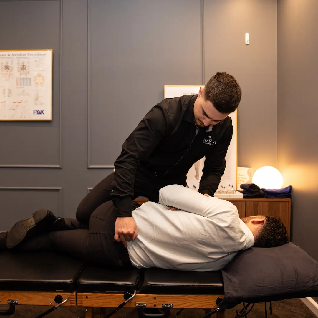 This is Dr Arthur at Aura Chiropractic performing a lower back Gonstead adjustment. Dr Arthur is wearing a black shirt with the aura chiropractic logo on it. The room is blue and the table is oak and black colour.