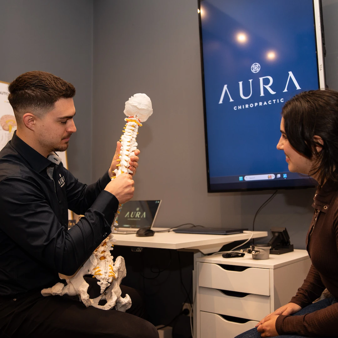 This visualises Dr Arthur Kaganovitch explaining an xray and spine to his female patient. They are both looking at the spinal model with intent. In the background, there is a Aura Chiropractic logo